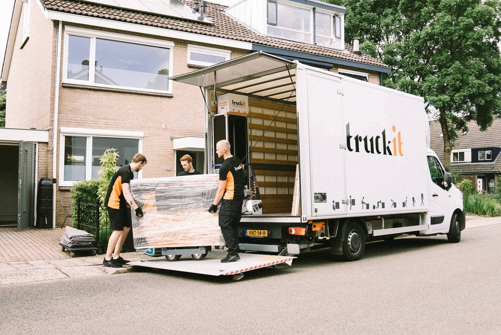 Drie verhuizers van Truckit rijden een ingepakte piano op de pianokar naar de laadklep van de verhuiswagen