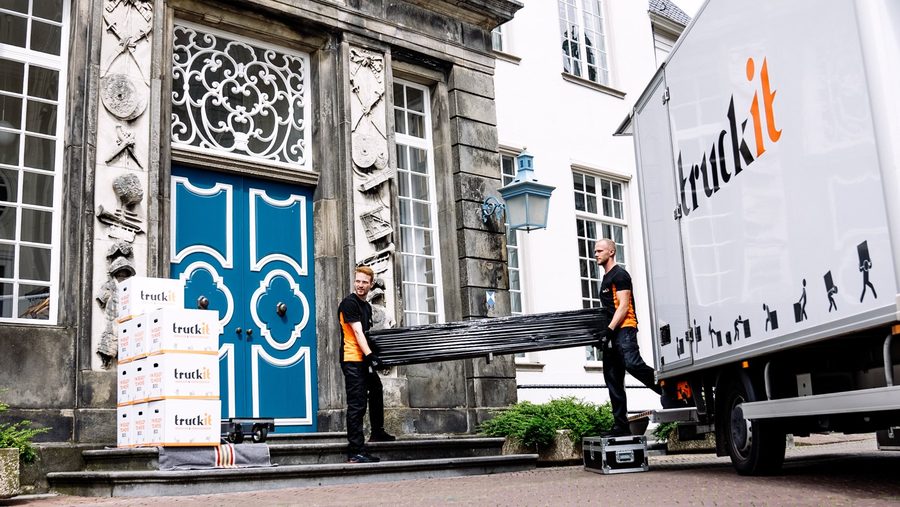Twee verhuizers van Truckit dragen een ingepakt meubel langs een monumentale voordeur naar de wagen