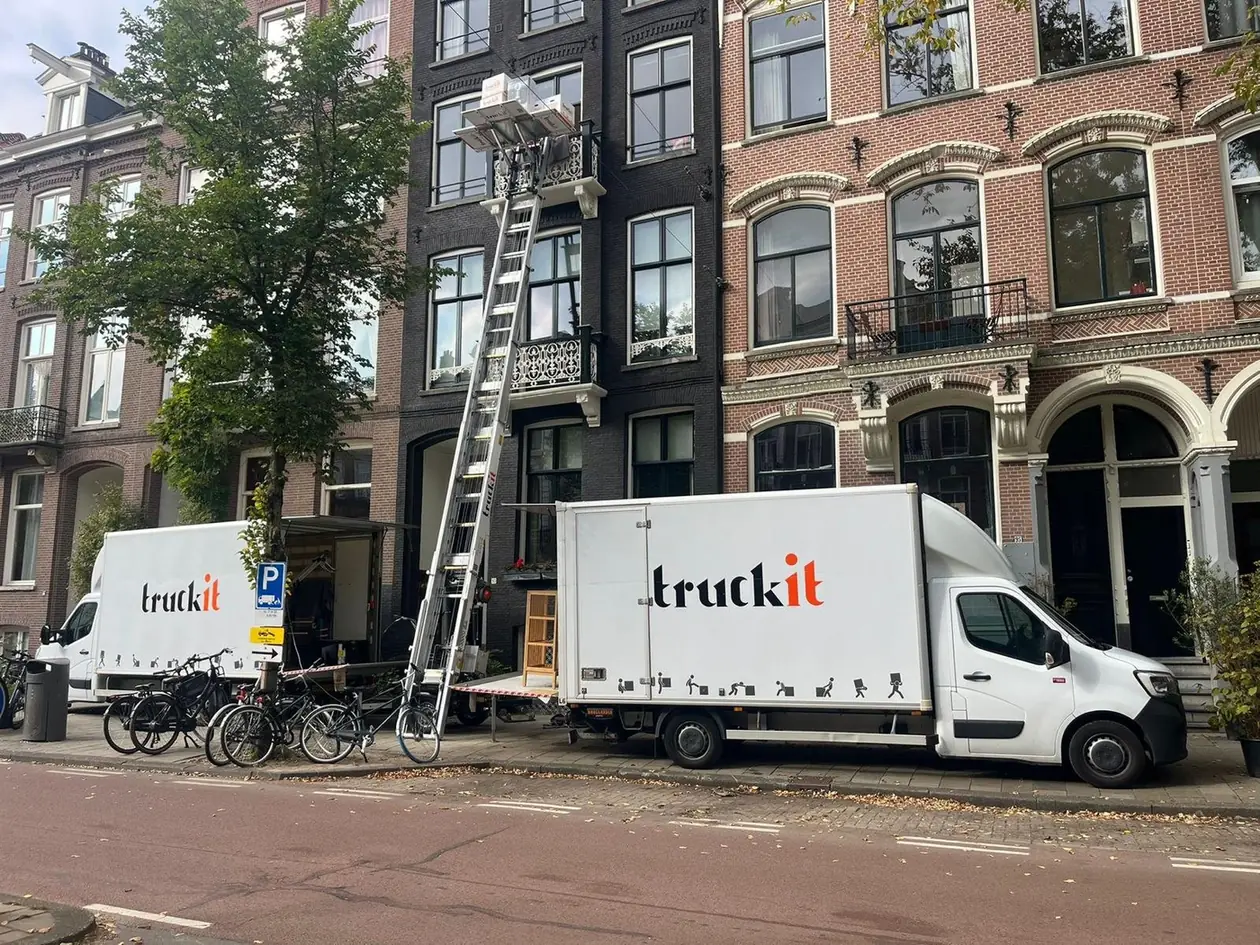 De verhuislift van Truckit tegen een grachtenpand in Amsterdam, met verhuisdozen op het plateau en twee verhuiswagens ervoor